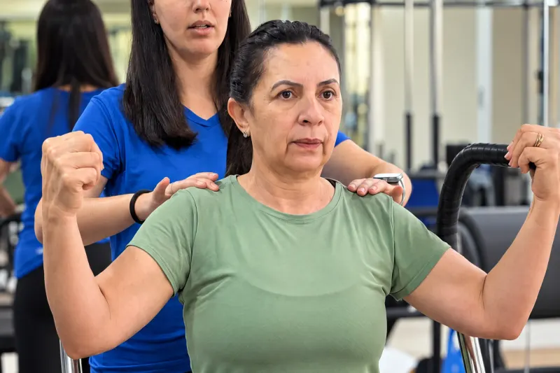 Professora da 2A ajusta os ombros de uma aluna na chair de Pilates